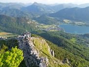 A group of hikers stands around a summit cross on a mountain, panoramic view of lake, mountains, sunny weather