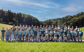 Large group of Eurofun team members in matching turquoise shirts standing on a green meadow with forest and blue sky in the background.