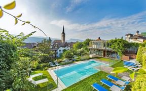 Outdoor swimming pool at Hotel Goldener Stern in Kaltern with blue loungers, surrounded by green lawn and trees. Church tower visible in background.