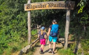 Adult and child walking through wooden entrance gate with 'Algunder Waalweg' sign, surrounded by lush green vegetation on hiking trail.
