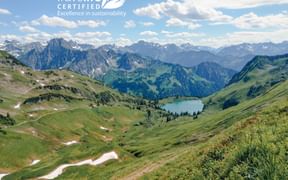 Panoramablick auf die Lechtaler Alpen mit Travelife-Zertifizierungslogo. Grüne Täler, Bergsee und schneebedeckte Gipfel unter bewölktem Himmel.
