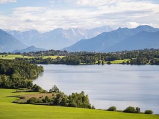 Malerischer Blick auf einen See umgeben von grünen Wiesen und Wäldern mit schneebedeckten Alpengipfeln im Hintergrund unter bewölktem Himmel.