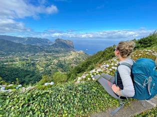 Female hiker with blue backpack sitting among white hydrangea flowers, overlooking Madeira's dramatic north coast landscape with mountains and ocean.