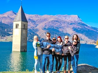 Sechs Menschen heben einen Mann in die Höhe vor dem Reschensee bei Sonnenschein