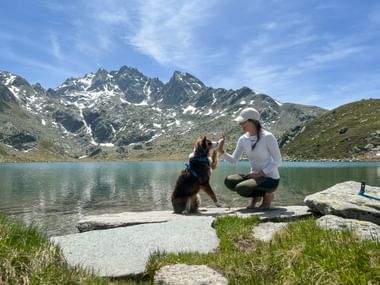 Frau in weißem Shirt und Kappe hockt neben Hund am felsigen Ufer des Surettasees. Schneebedeckte Berge und blauer Himmel im Hintergrund.