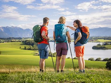 Three hikers on a meadow with Lake Riegsee in the background