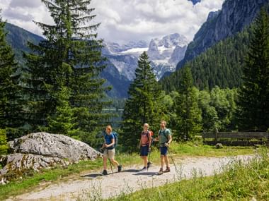 Drei Wanderer mit Rucksäcken auf einem Bergweg beim Gosausee mit schneebedeckten Gipfeln, grünen Wäldern und einem See im Hintergrund.