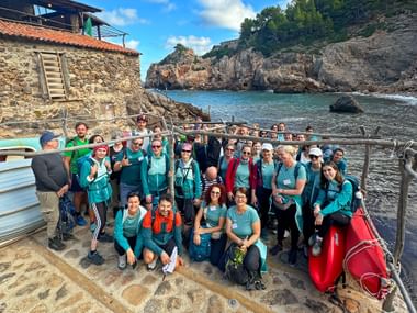 Large group of hikers in matching turquoise shirts posing at a rocky coastal cove in Mallorca with stone buildings and cliffs in the background.
