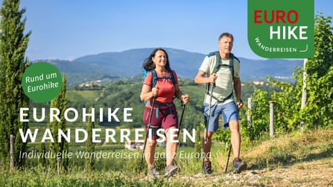 Zwei Wanderer mit Rucksäcken und Wanderstöcken gehen durch Weinbergreihen mit Hügeln und Bergen im Hintergrund. Eurohike-Logo sichtbar.